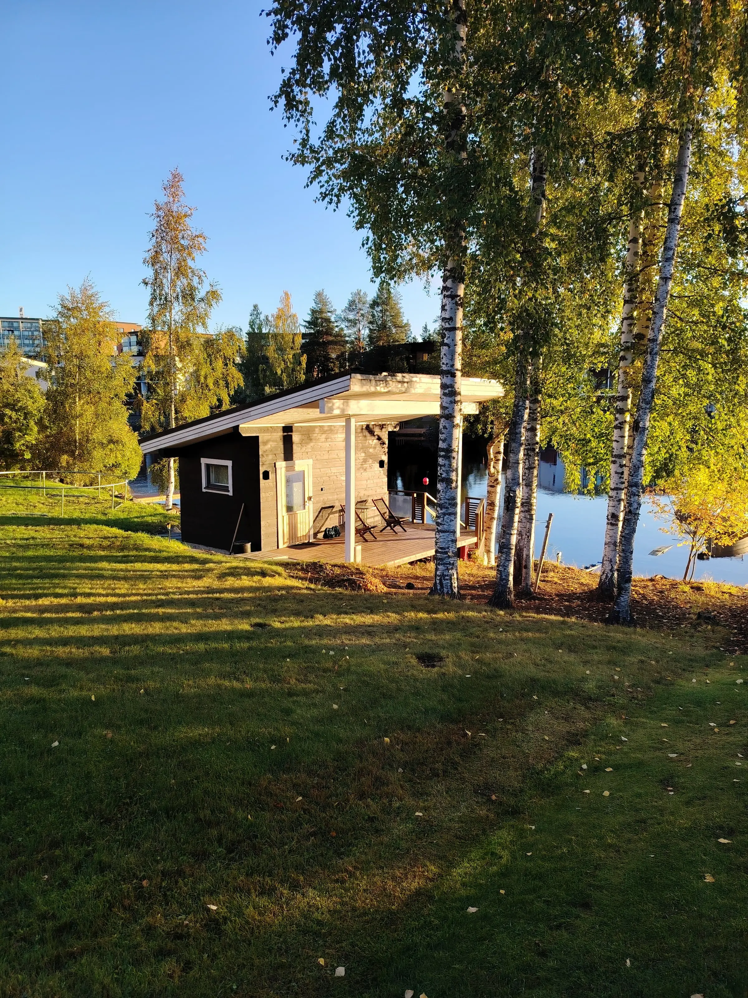 Rantasauna järven rannalla nurmikon yli nähtynä, koivut ja järvi oikealla.