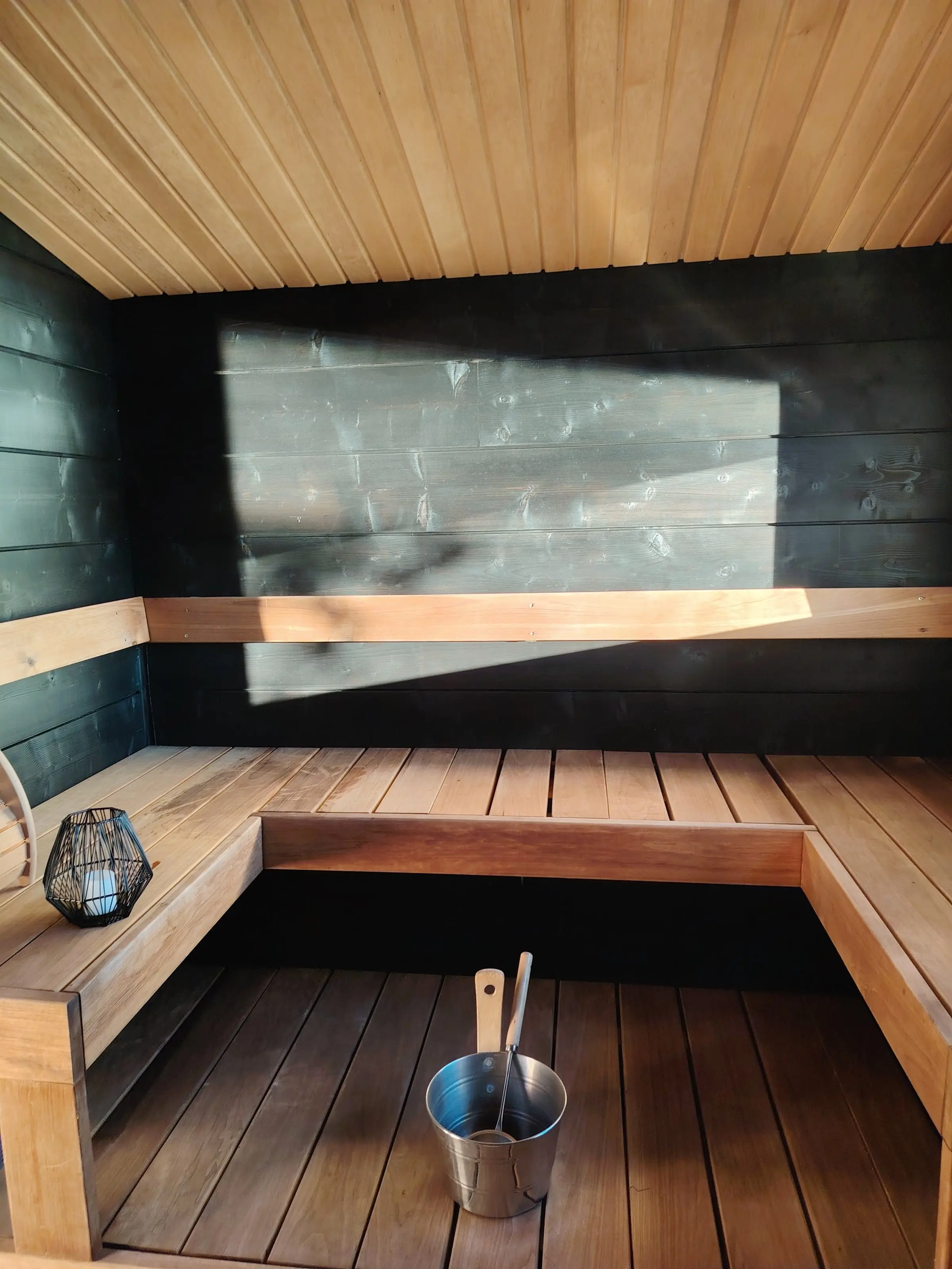 Morning light on the dark walls and L-shaped benches of the sauna room, with a lantern and a sauna bucket.